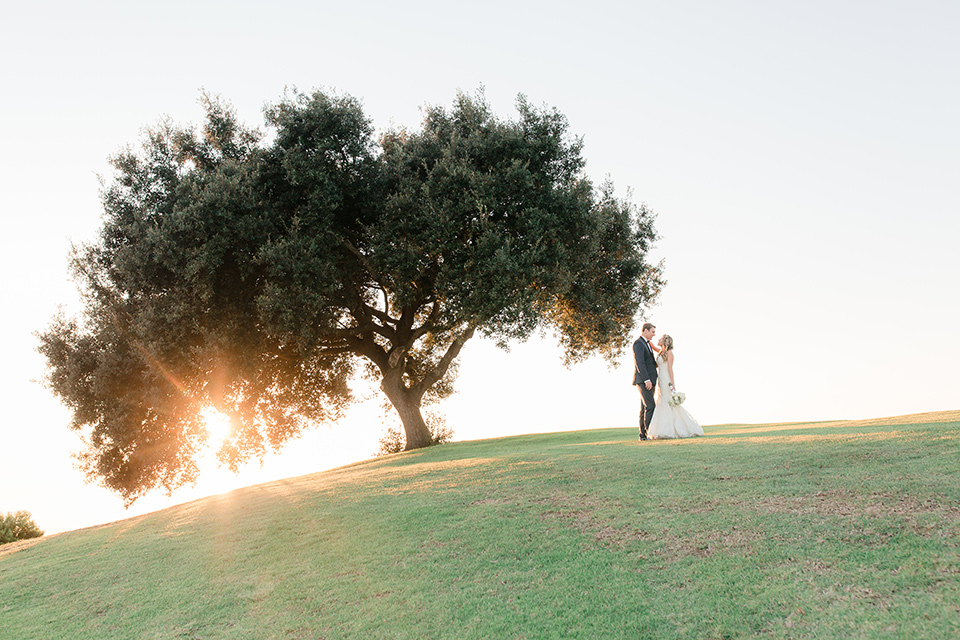 Los-Verdes-Golf-Course-Wedding-bride-and-groom-near-giant-tree-bride-is-in-a-lace-mermaid-style-gown-with-a-modified-sweetheart-neckline-the-groom-is-in-a-navy-blue-tuxedo-with-a-black-shawl-lapel-and-a-black-bow-tie