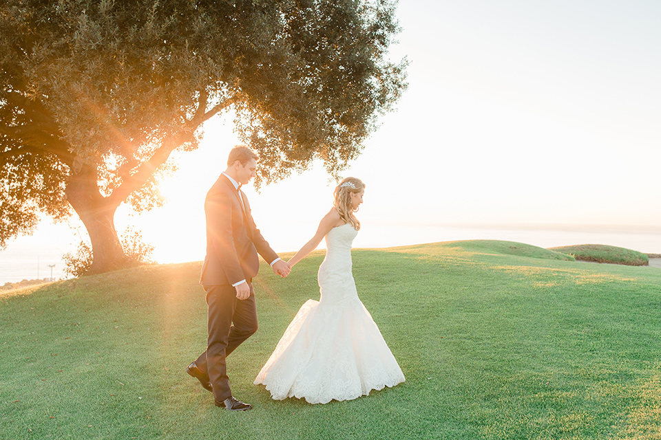 Los-Verdes-Golf-Course-Wedding-bride-and-groom-walking-by-tree-bride-is-in-a-lace-mermaid-style-gown-with-a-modified-sweetheart-neckline-the-groom-is-in-a-navy-blue-tuxedo-with-a-black-shawl-lapel-and-a-black-bow-tie