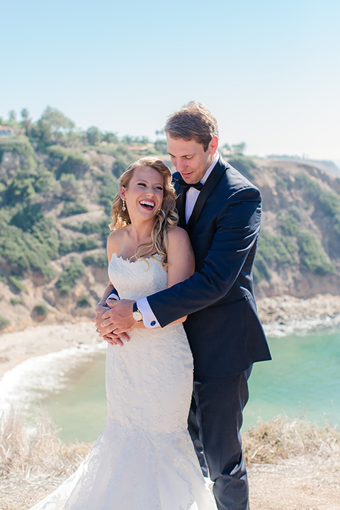 Los-Verdes-Golf-Course-Wedding-groom-holding-bride-from-behind-bride-is-in-a-lace-mermaid-gown-with-a-modified-sweetheart-neckline-and-the-groom-is-in-a-navy-blue-shawl-lapel-tuxedo