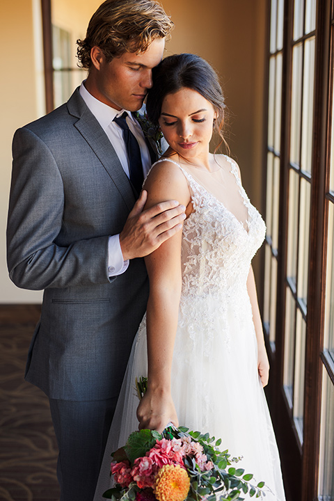 aliso Viejo wedding design with the bride and groom by the windows the bride is in a lace gown with thin straps and the groom in a grey suit with a navy neck tie