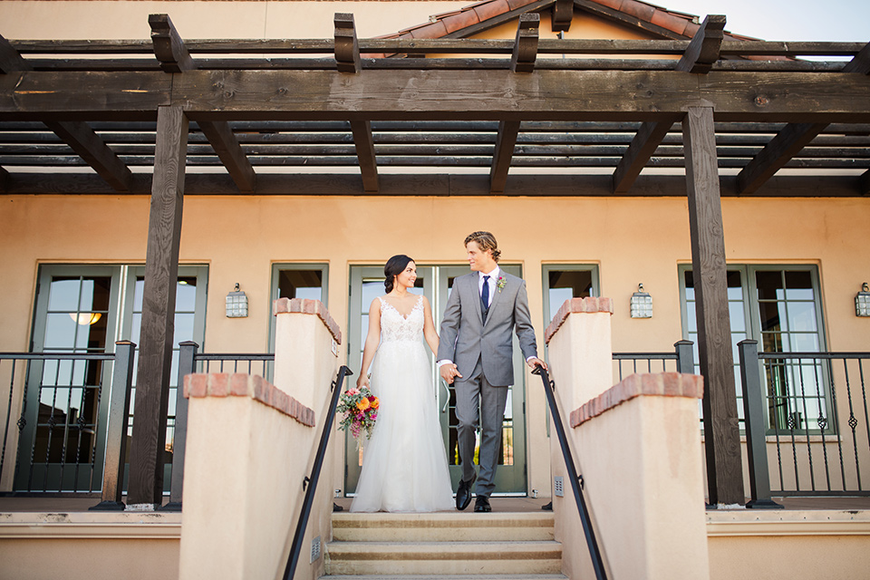 Aliso Viejo wedding design with the bride  and groom at the top of the stairs the bride in a lace gown with thin straps and the groom in a grey suit with a blue neck tie