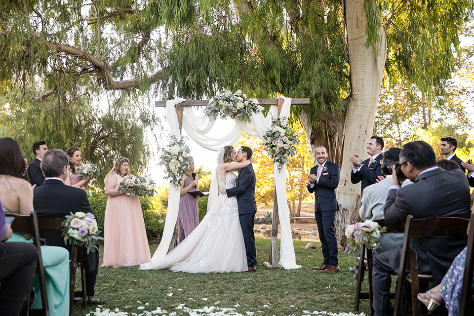 Garden-Wedding-first-kiss-bridesmaids-in-pink-long-dresses-groomsmen-are-in-navy-suits-with-pink-vests-and-pink-bow-ties-bride-in-a-strapless-ballgown-and-groom-in-a-navy-suit-with-a-white-long-tie