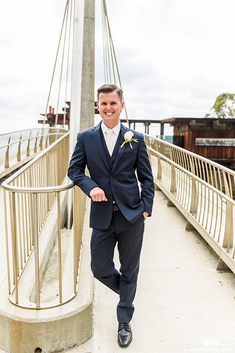 Groom leaning against railing in a navy blue suit and white tie