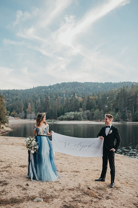 Big-Bear-Elopement-Shoot-bride-and-groom-holding-just-married-sign-bride-in-a-blue-tulle-gown-with-an-illusion-neckline-and-floral-design-the-groom-wore-a-traditional-black-tuxedo-with-a-peak-lapel-and-black-bow-tie