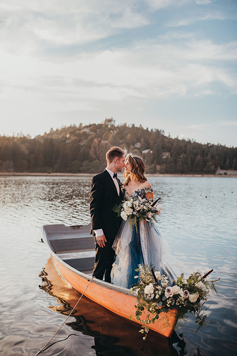 Big-Bear-Elopement-Shoot-bride-and-groom-standing-in-boat-bride-is-in-a-blue-tulle-gown-with-an-illusion-neckline-and-floral-design-the-groom-wore-a-traditional-black-tuxedo-with-a-peak-lapel-and-black-bow-tie