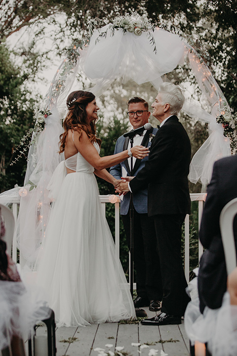 West-Hollywood-Wedding-brides-holding-hands-at-ceremony-one-bride-in-a-black-tuxedo-with-a-grey-cvest-and-black-bow-tie-the-other-bride-in-a-flowing-white-gowin-with-a-sweetheasrt-neckline-and-white-straps