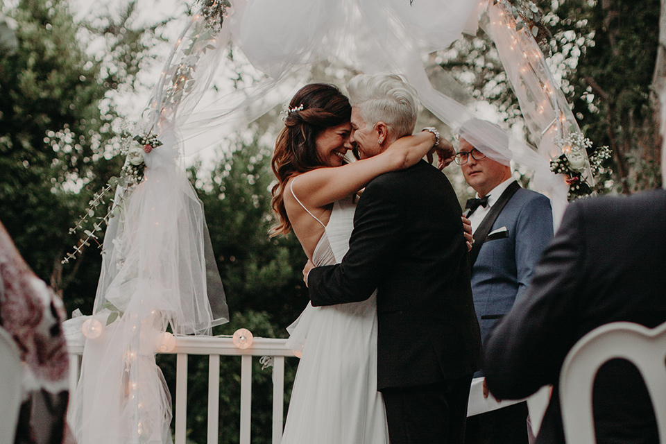 West-Hollywood-Wedding-brides-hugging-at-ceremony-one-bride-in-a-black-tuxedo-with-a-grey-vest-and-black-bow-tie-the-other-bride-in-a-flowing-white-gowin-with-a-sweetheasrt-neckline-and-white-straps