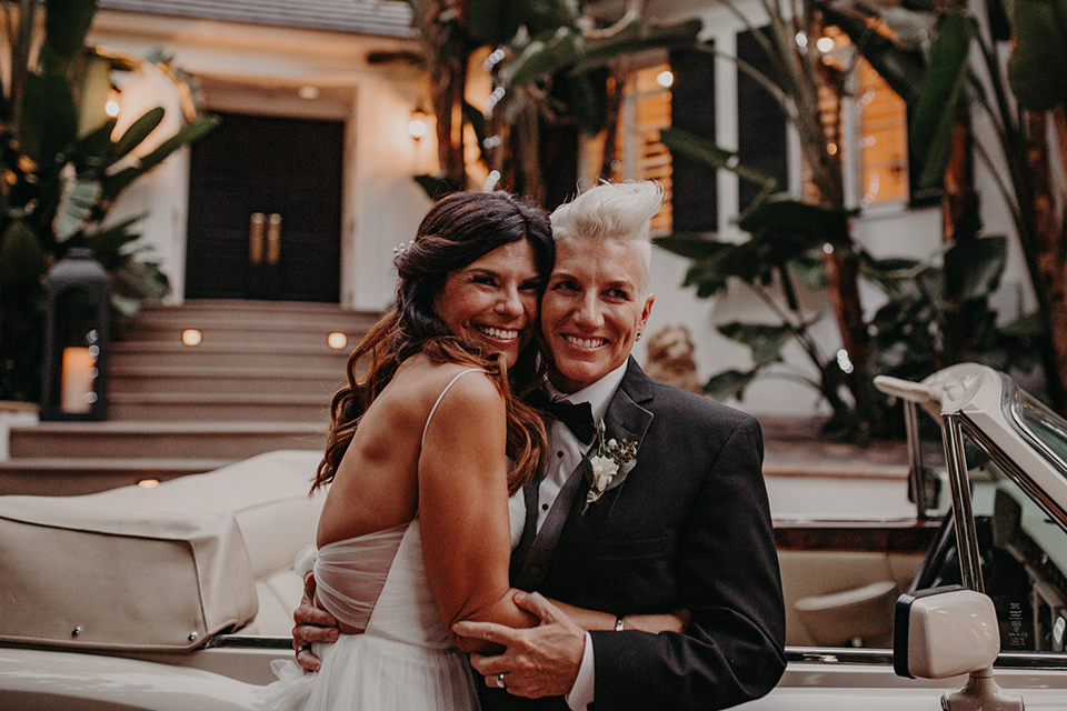 West-Hollywood-Wedding-brides-huging-by-car-one-bride-in-a-black-tuxedo-with-a-grey-vest-and-black-bow-tie-the-other-bride-in-a-flowing-white-gowin-with-a-sweetheasrt-neckline-and-white-straps