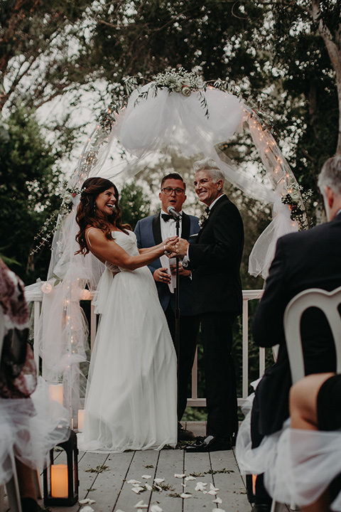 West-Hollywood-Wedding-brides-laughing-at-ceremony-one-bride-in-a-black-tuxedo-with-a-grey-vest-and-black-bow-tie-the-other-bride-in-a-flowing-white-gowin-with-a-sweetheasrt-neckline-and-white-straps