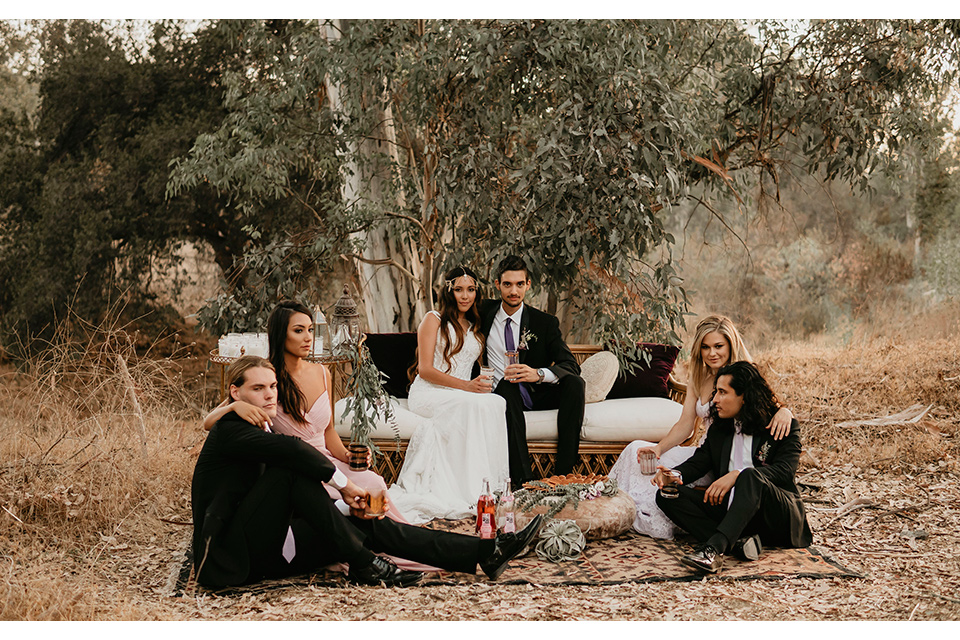 Boho chic elopement wedding with the bridesmaids with soft pink gowns and the groomsmen in black suits the bride in an ivory lace ball gown with a gold chain headpiece and the groom in a black suit with a black long tie and everyone is sitting on the ground with bohemian pillows and carpet