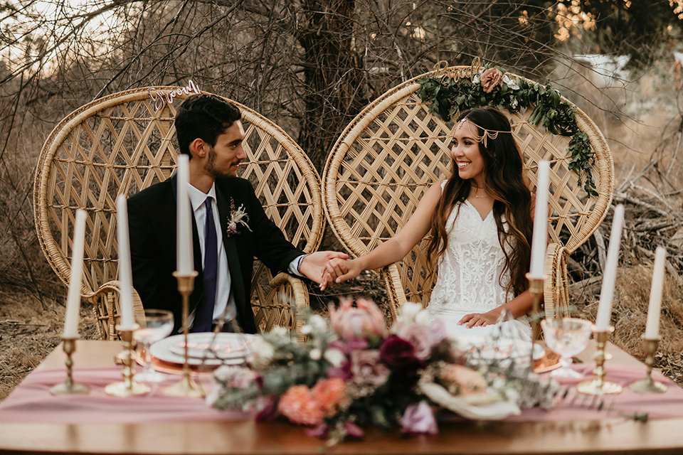 Boho chic elopement wedding with the bride in an ivory lace ball gown with a gold chain headpiece and the groom in a black suit with a black long tie sitting at the sweetheart table in big wicker chairs