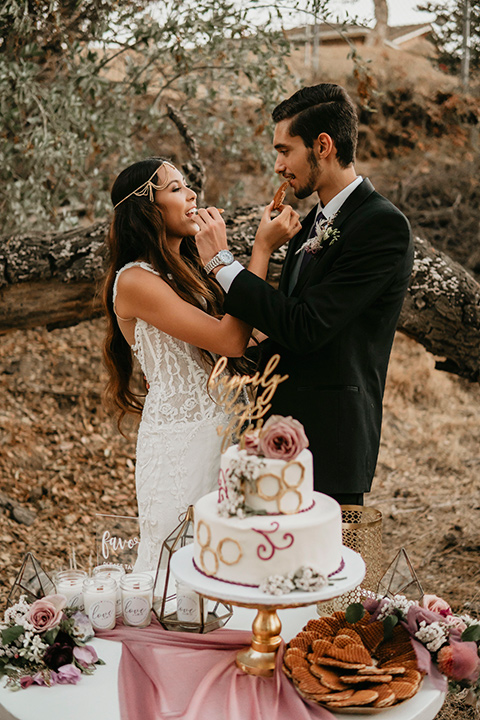 boho elopement shoot with the bride in a lace ball gown and a gold chair headpiece and the groom in a black suit with a long black tie cutting the cake