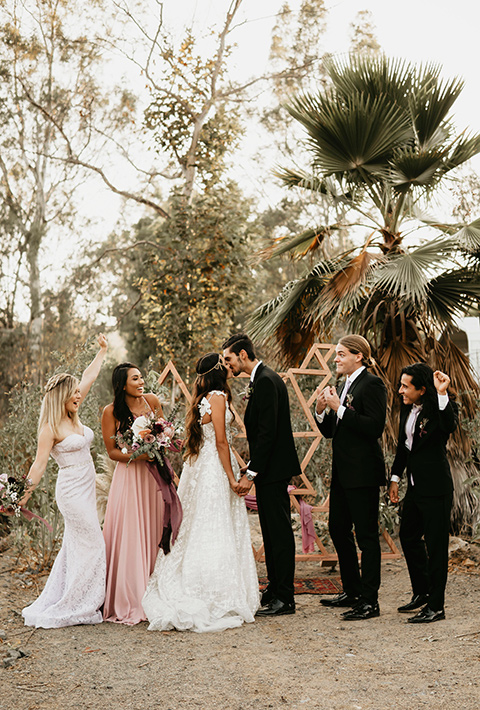 bride in an ivory lace gown with a gold chain headpiece and the groom in a black suit with a black long tie and the bridesmaids in pink dresses with the groomsmen in black suits and black ties