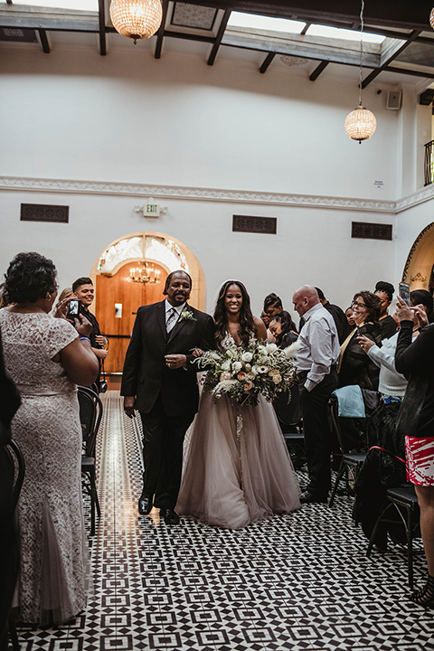 The-Ebell-In-Long-Beach-Wedding-bride-walking-down-the-aisle-bride-in-a-champagne-ballgown-with-a-strapless-neckline-and-the-groom-in-a-black-tuxedo-with-a-rose-gold-bow-tie