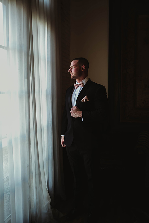 The-Ebell-In-Long-Beach-Wedding-groom-looking-out-the-window-in-a-black-tuxedo-with-a-rose-gold-bow-tie