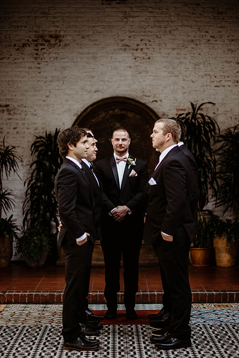 The-Ebell-In-Long-Beach-Wedding-groomsmen-standing-facing-each-other-groom-in-the-middle-close-up-groom-in-a-black-tuxedo-with-a-rose-gold-bow-tie-groomsmen-in-black-tuxedos-with-black-long-ties