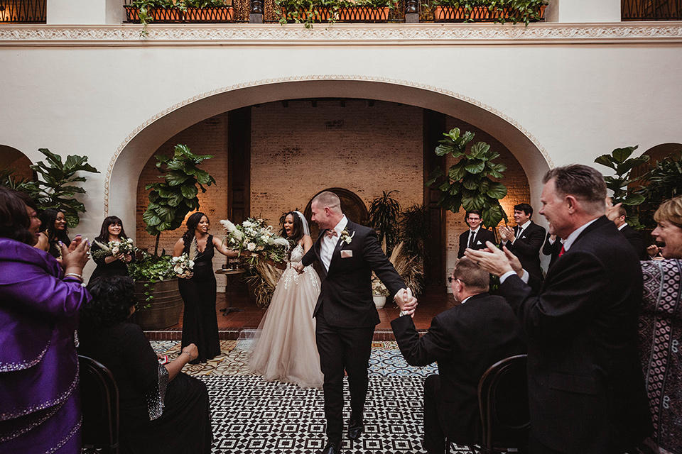 The-Ebell-In-Long-Beach-Wedding-walking-down-the-aisle-bride-in-a-champagne-ballgown-with-a-strapless-neckline-and-the-groom-in-a-black-tuxedo-with-a-rose-gold-bow-tie