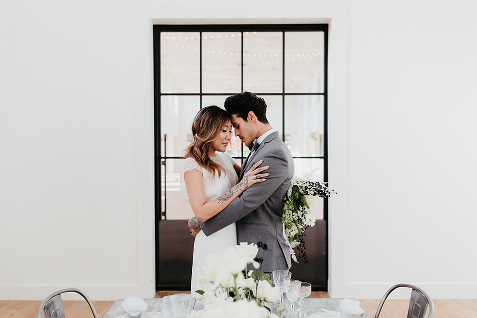 Building-177-Styled-Shoot-bride-and-groom-by-table-touching-heads-bride-in-a-flowing-white-gown-with-short-sleeves-and-beading-detail-groom-in-a-grey-suit-with-a-grey-velvet-bow-tie