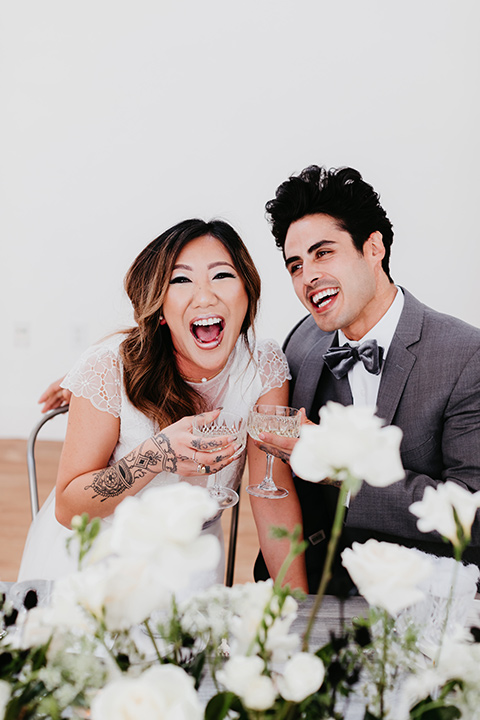 Building-177-Styled-Shoot-bride-and-groom-with-drinks-bride-in-a-flowing-white-gown-with-short-sleeves-and-beading-detail-groom-in-a-grey-suit-with-a-grey-velvet-bow-tie