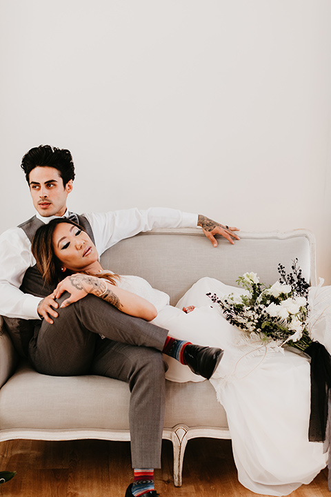 Building-177-Styled-Shoot-bride-and-groom-laying-on-couch-bride-in-a-flowing-white-gown-with-short-sleeves-and-beading-detail-groom-in-a-grey-suit-with-a-grey-velvet-bow-tie