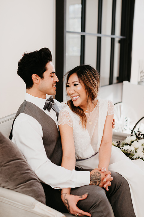 Building-177-Styled-Shoot-bride-and-groom-sitting-on-couch-bride-in-a-flowing-white-gown-with-short-sleeves-and-beading-detail-groom-in-a-grey-suit-with-a-grey-velvet-bow-tie