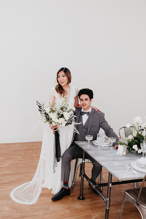 Building-177-Styled-Shoot-groom-sitting-and-bride-standing-by-table-bride-in-a-flowing-white-gown-with-short-sleeves-and-beading-detail-groom-in-a-grey-suit-with-a-grey-velvet-bow-tie