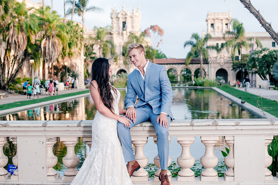 California  Dreaming  Shoot  bride  and  groom  sitting  on  ledge  bride  in  a  lace  form  fitting gown with  thin straps groom in  a  light  blue  suit