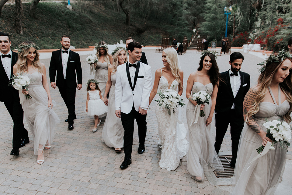 rancho-las-lomas-real-wedding-bridalparty-walking-the-bridesmaids-in-nude-and-taupe-long-gowns-the-groomsmen-in-black-tuxedos-with-black-bow-ties-the-bride-is-in-a-lace-long-gown-with-a-deep-v-and-straps-and-the-groom-is-in-a-white-shawl-lapel-tuxedo-with-black-pants-and-a-black-bow-tie