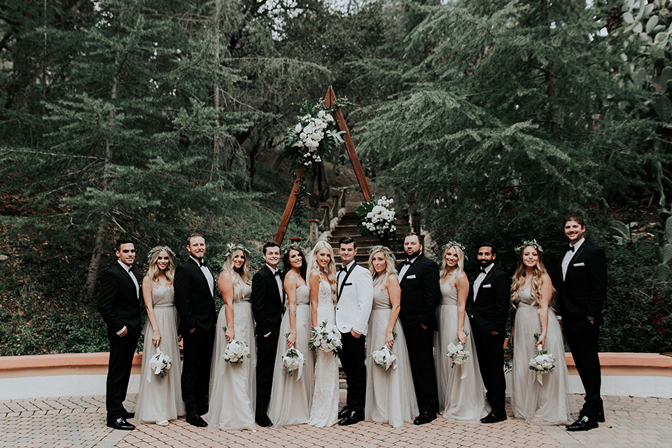 rancho-las-lomas-real-wedding-bridalparty-the-bridesmaids-in-nude-and-taupe-long-gowns-the-groomsmen-in-black-tuxedos-with-black-bow-ties-the-bride-is-in-a-lace-long-gown-with-a-deep-v-and-straps-and-the-groom-is-in-a-white-shawl-lapel-tuxedo-with-black-pants-and-a-black-bow-tie