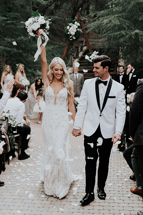 rancho-las-lomas-real-wedding-bride-and-groom-walking-down-the-aisle-the-bride-in-a-lace-white-gown-with-a-straps-and-a-deep-v-neckline-the-groom-in-a-white-shawl-lapel-tuxedo-with-black-pants-and-a-black-bow-tie