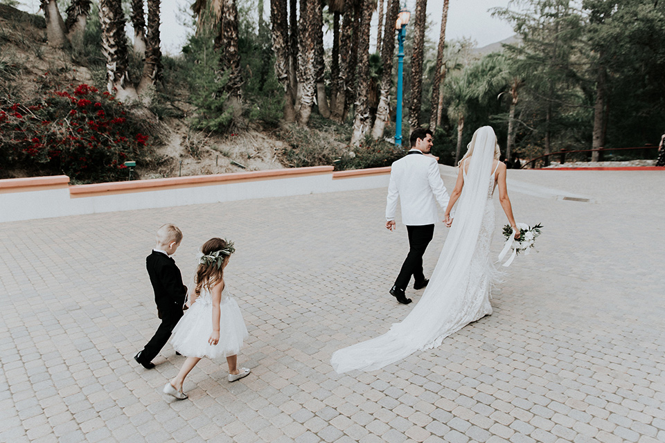 rancho-las-lomas-real-wedding-bride-and-groom-walking-with-children-the-bride-is-in-a-lace-long-gown-with-a-deep-v-and-straps-and-the-groom-is-in-a-white-shawl-lapel-tuxedo-with-black-pants-and-a-black-bow-tie