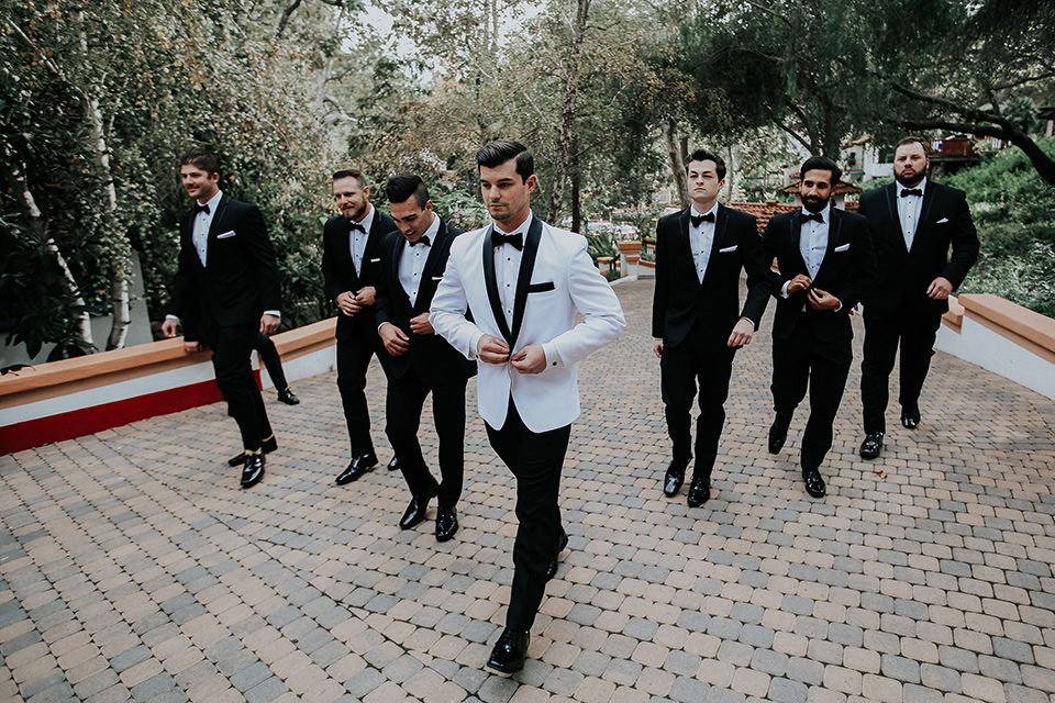 rancho-las-lomas-real-wedding-groom-and-groomsmen-walking-buttoning-jacket-the-groomsmen-in-black-tuxedos-with-black-bow-ties-the-groom-is-in-a-white-shawl-lapel-tuxedo-with-black-pants-and-a-black-bow-tie