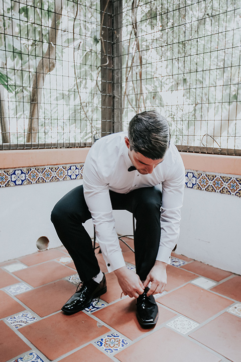 rancho-las-lomas-real-wedding-groom-tying-shoe-in-a-white-shawl-lapel-tuxedo-with-black-pants-and-a-black-bow-tie