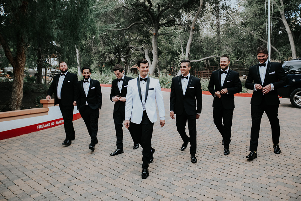 rancho-las-lomas-real-wedding-groomsmen-walking-the-groomsmen-in-black-tuxedos-with-black-bow-ties-the-groom-is-in-a-white-shawl-lapel-tuxedo-with-black-pants-and-a-black-bow-tie