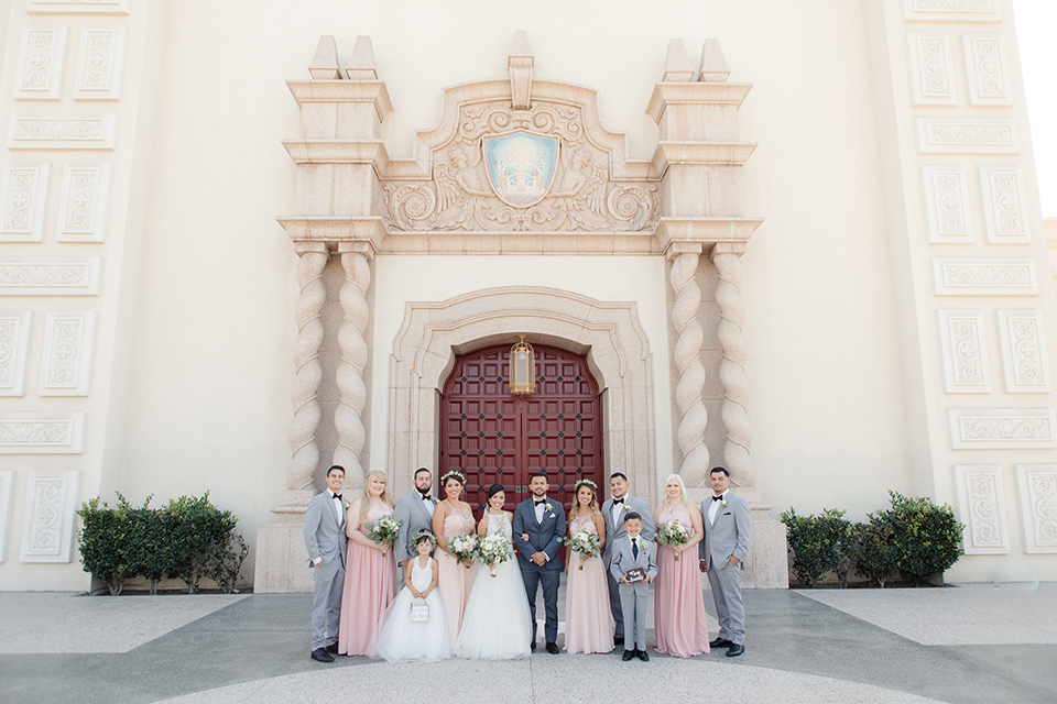 kellogg-house-pomona-wedding-bridal-party-outside-venue-the-bridesmaids-are-in-pink-toned-gowns-the-groomsmen-in-light-grey-suits-bride-in-a-mermaid-style-gown-with-a-high-neckline-and-the-groom-in-a-dark-grey-tuxedo