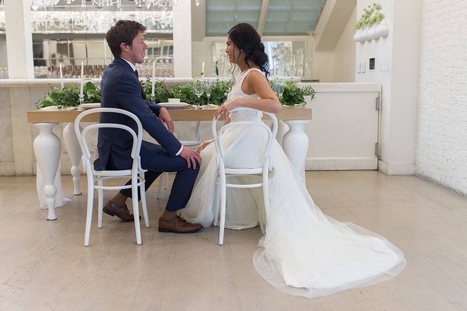 Cooks-Chapel-Shoot-bride-and-groom-sitting-bride-is-in-a-white-flowing-gown-with-a-deep-v-neckline-groom-is-in-a-dark-blue-suit-with-a-navy-long-tie-and-brown-shoes