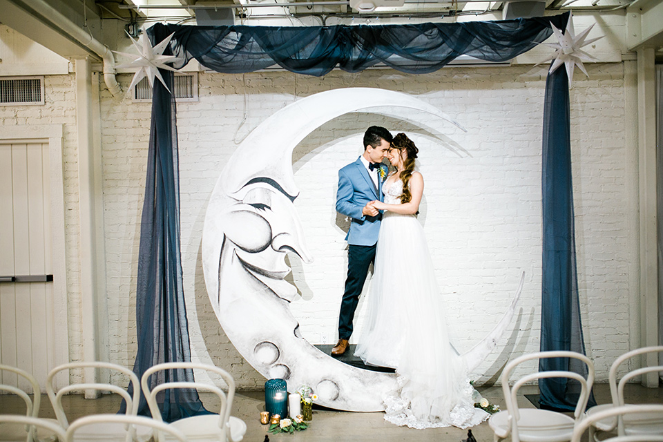 Wedding bride and groom at moon ceremony arch with the bride in a white ballgown with a lace bodice and straps. The groom is in a light blue coat with a dark blue vest and pants with a dark blue bow tie