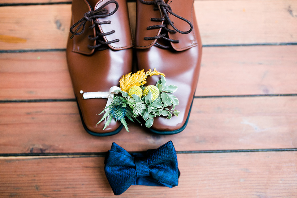 Groom's wedding shoe and accessories and a navy bow tie on top of a wood background