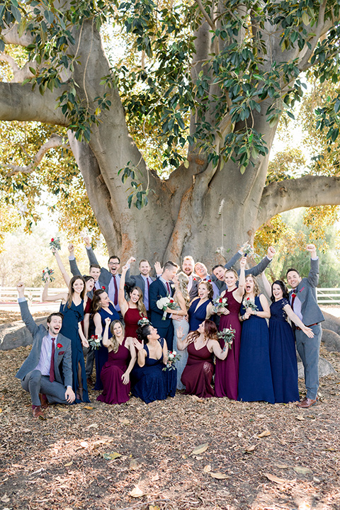 red-barn-wedding-bridal-party-hands-in-the-air-bridesmaids-in-alternating-navy-and-burgundy-dresses-groomsmen-in-grey-suits-bride-in-a-lace-fitted-dress-with-thin-straps-groom-in-a-blue-suit-with-grey-vest-and-burgundy-tie