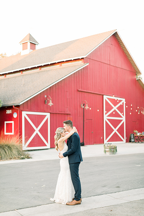 red-barn-wedding-bride-and-groom-in-front-of-barn-bride-in-a-lace-fitted-dress-with-thin-straps-groom-in-a-blue-suit-with-grey-vest-and-burgundy-tie