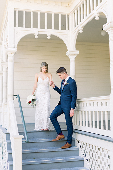 red-barn-wedding-bride-and-groom-walking-down-steps-of-white-building-bride-in-a-lace-fitted-dress-with-thin-straps-groom-in-a-blue-suit-with-grey-vest-and-burgundy-tie