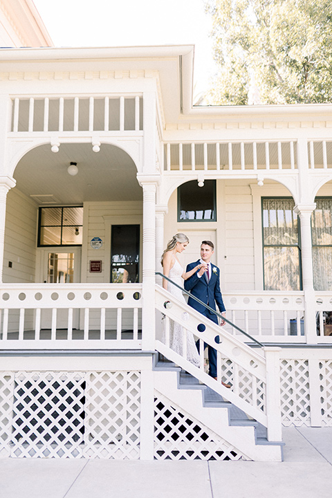 red-barn-wedding-bride-and-groom-walking-down-steps-of-white-building-bride-in-a-lace-fitted-dress-with-thin-straps-groom-in-a-blue-suit-with-grey-vest-and-burgundy-tie