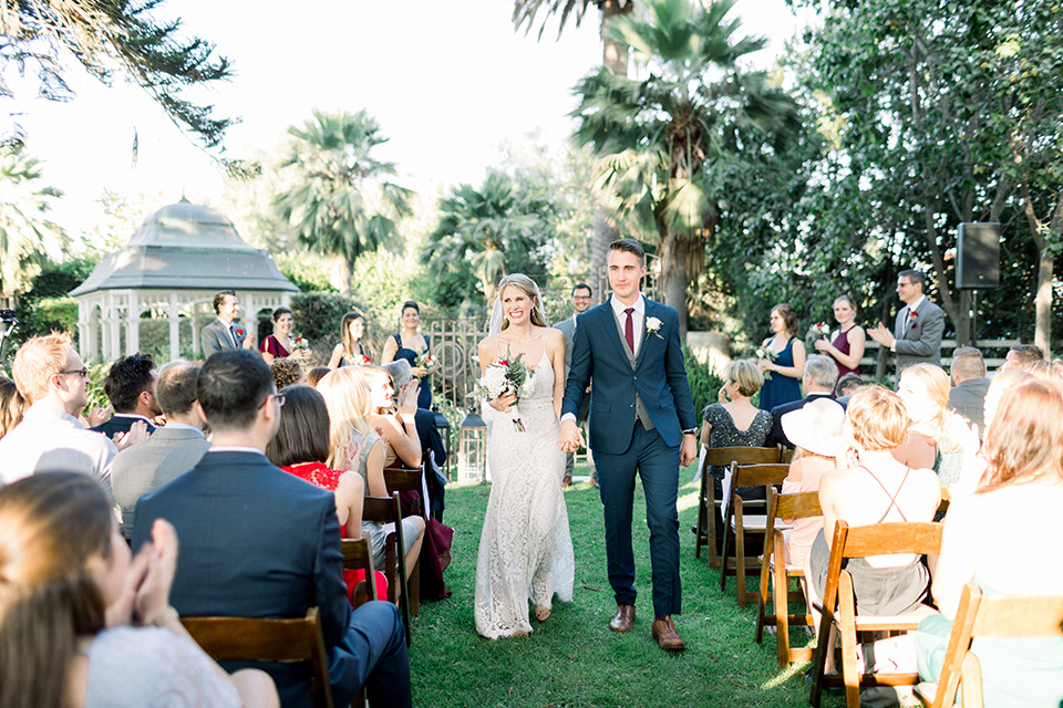 red-barn-wedding-bride-and-groom-walking-down-after-ceremont-bride-in-a-lace-fitted-dress-with-thin-straps-groom-in-a-blue-suit-with-grey-vest-and-burgundy-tie