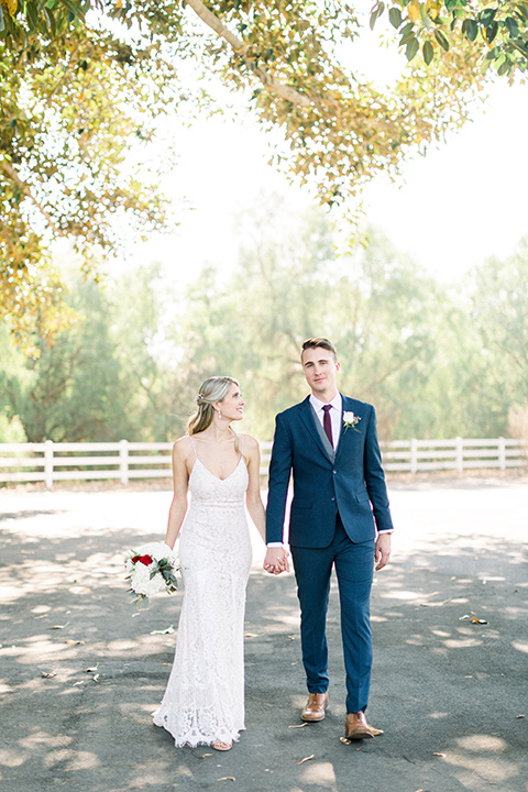 red-barn-wedding-bride-and-groom-walking-bride-in-a-lace-fitted-dress-with-thin-straps-groom-in-a-blue-suit-with-grey-vest-and-burgundy-tie