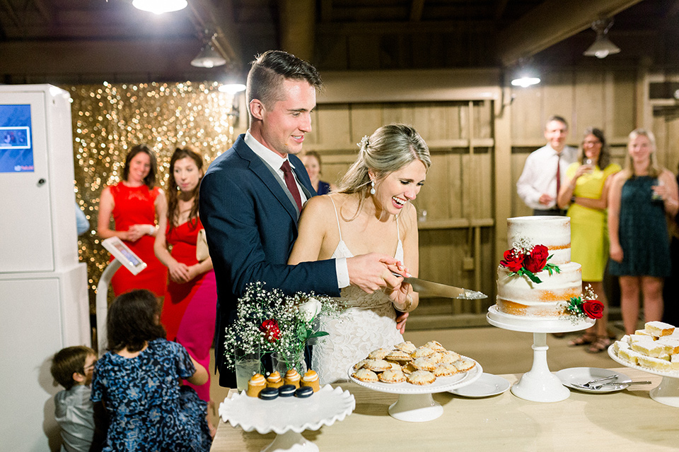 red-barn-wedding-cutting-the-cake-bride-in-a-lace-fitted-dress-with-thin-straps-groom-in-a-blue-suit-with-grey-vest-and-burgundy-tie