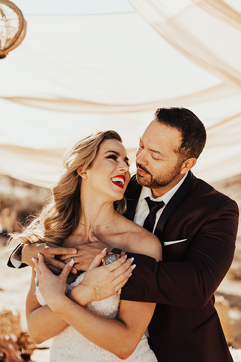 Bohemian-Desert-Shoot-bride-and-groom-holding-each-other-bride-in-a-formfitting-lace-gown-with-a-strapless-neckline-groom-in-a-burgundy-shawl-lapel-tuxedo-with-a-black-long-lie