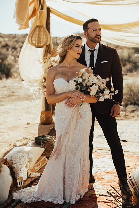 Bohemian-Desert-Shoot-bride-and-groom-looking-away-from-camera-bride-in-a-lace-formfitting-gown-with-a-sweetheart-neckline-groom-in-a-burgundy-shawl-lapel-tuxedo-with-a-black-long-tie