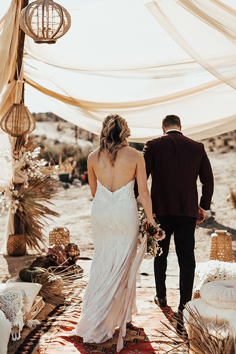 Bohemian-Desert-Shoot-bride-and-groom-moving-away-from-camera-bride-in-a-lace-formfitting-gown-with-a-sweetheart-neckline-groom-in-a-burgundy-shawl-lapel-tuxedo-with-a-black-long-tie