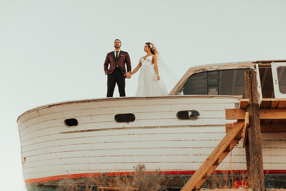 Bohemian-Desert-Shoot-bride-and-groom-on-boat-bride-in-a-formfitting-gown-with-lace-detailing-and-a-sweetheart-neckline-groom-in-a-burgundy-shawl-lapel-tuxedo-with-a-black-long-tie