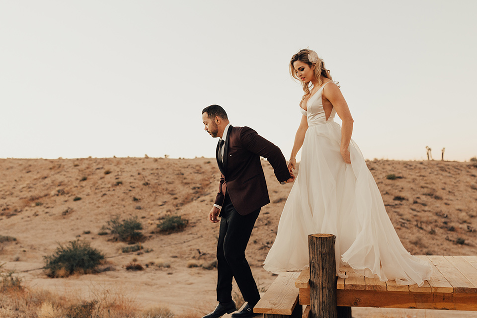 Bohemian-Desert-Shoot-bride-and-groom-walking-down-steps-bride-in-a-formfitting-gown-with-lace-detailing-and-a-sweetheart-neckline-groom-in-a-burgundy-shawl-lapel-tuxedo-with-a-black-long-tie